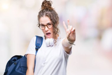 Mutlu yüz zafer işareti yaparken kameraya göz kırpıyor ile gülümseyen izole arka plan üzerinde sırt çantası ve kulaklıklar giyen genç esmer öğrenci kız. İki numaralı.
