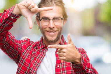 Genç yakışıklı adam uzun saçlı gözlüklü elle yapma çerçeve ve parmakları ile mutlu yüz gülümseyen izole arka plan üzerinde. Yaratıcılık ve Fotoğrafçılık kavramı.