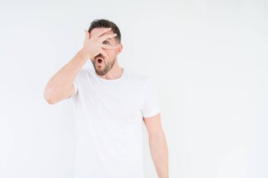 Mahcup ifadeyle parmaklarımın arasından seyir yüz ve gözler el ile kapsayan şok bakmak izole arka plan üzerinde rahat beyaz t-shirt giyen genç yakışıklı adam.