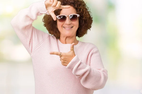 美しい中間エイガー年配の女性の笑顔の幸せそうな顔で指と手作りフレーム分離の背景にピンクのセーター サングラスを着用します 創造性と写真のコンセプト — ストック写真