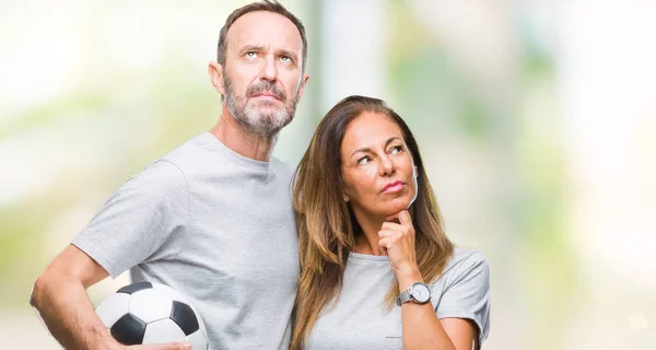 Middle age hispanic couple holding football soccer ball over isolated background serious face thinking about question, very confused idea
