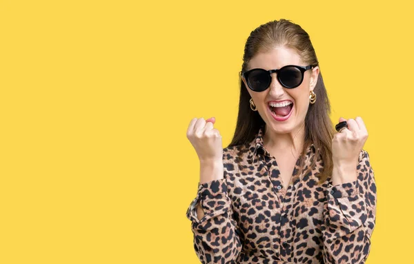 Middle age mature rich woman wearing sunglasses and leopard dress over isolated background celebrating surprised and amazed for success with arms raised and open eyes. Winner concept.