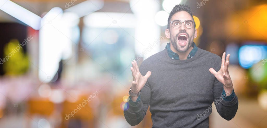 Young handsome man wearing glasses over isolated background crazy and mad shouting and yelling with aggressive expression and arms raised. Frustration concept.