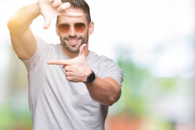 Genç yakışıklı adam elleriyle yapım çerçeve ve parmakları ile mutlu yüz gülümseyen izole arka plan üzerinde güneş gözlüğü takıyor. Yaratıcılık ve Fotoğrafçılık kavramı.