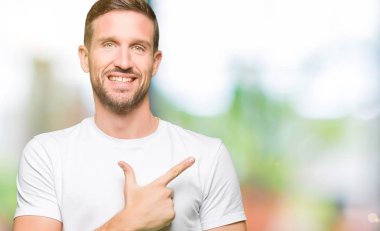 Yakışıklı adam neşeli bir gülümseme ile el ve parmak kadar yan yüzünde mutlu ve doğal ifade ile işaret yüz ile rahat beyaz t-shirt giyen