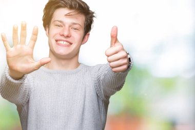 Gösterilen arka plan üzerinde kış kazak giyen genç yakışıklı adam izole ve parmakları ile işaret kendinden emin ve mutlu gülümseyerek süre 6 numara.