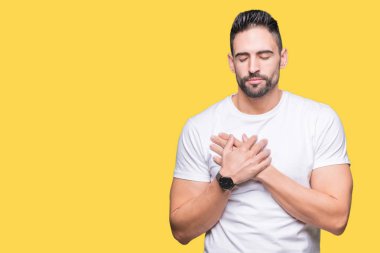 Genç adam ile gülümseyen izole arka plan üzerinde rahat beyaz t-shirt giyen kapalı gözler ve yüzünde minnettar jest ile göğüs üzerinde eller. Sağlık kavramı.