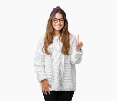Ve parmak gülümseyen süre bir numara ile kendinden emin ve mutlu yukarıyı gösteren izole arka plan üzerinde gözlük ve kış şapka giyen genç güzel esmer hipster kadın.