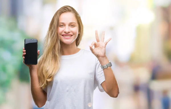 年轻美丽的金发碧眼的女人显示屏幕的智能手机在孤立的背景做确定的标志与手指 优秀的象征 — 图库照片