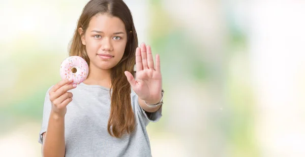 年轻的美丽的女人吃粉红色的甜甜圈在孤立的背景与开放的手做停止标志与严肃和自信的表情 防御姿态 — 图库照片