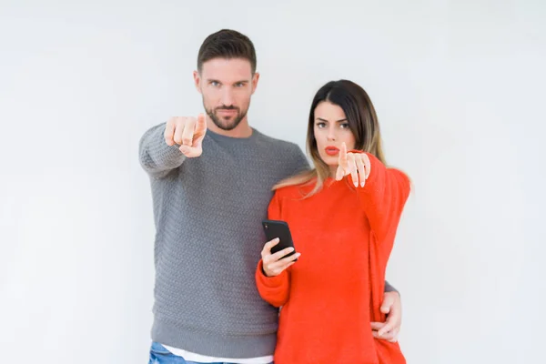 年轻夫妇使用智能手机在孤立的背景指向手指对着相机和你 手的标志 积极和自信的手势从前面 — 图库照片