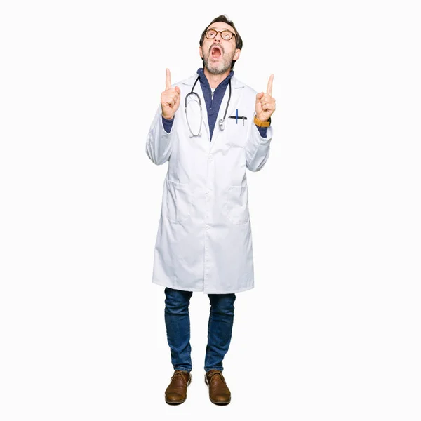 英俊的中年医生男子穿着医疗外套惊讶和惊讶地抬起头来 用手指和举起的手臂指指点点 — 图库照片
