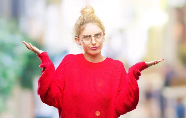 若い美しい金髪女性の腕と手を上げて分離背景無知と混乱する式で赤いセーターを着て 眼鏡を着用します 疑いのコンセプト — ストック写真