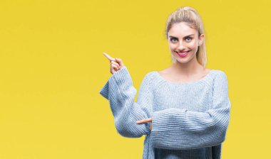 Yüzünde kocaman bir gülümseme ile izole arka plan üzerinde kış kazak giyen genç güzel sarışın ve mavi gözlü kadın kameraya bakarak yan el ve parmak ile işaret.