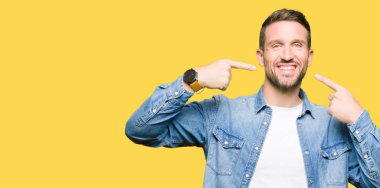 Kendinden emin gösterilen ve işaret parmakları diş ve ağız ile gülümseyen denim ceket giyen sakal ve mavi gözlü yakışıklı adam. Sağlık kavramı.