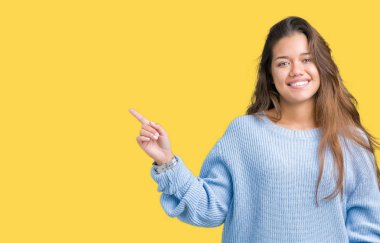 Kameraya bakarak yan el ve parmak ile işaret yüzünde kocaman bir gülümseme ile izole arka plan üzerinde mavi kış kazak giyen genç güzel esmer kadın.