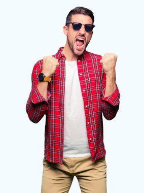 Gülümseyen ve başarı için çığlık çok mutlu ve heyecanlı kaldırdı, silah ile kazanan jest yapmak moda güneş gözlüğü takıyor yakışıklı adam. Kutlama kavramı.