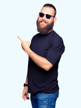 Yüzünde, kocaman bir gülümseme ile güneş gözlüğü takmış genç hipster adam kameraya bakarak yan el ve parmak ile işaret.