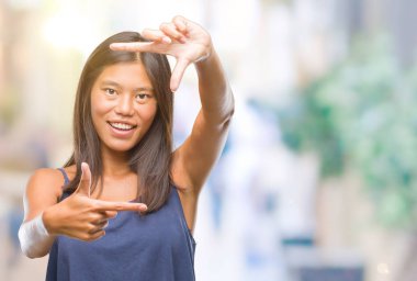 Genç Asyalı kadın elle yapma çerçeve ve parmakları ile mutlu yüz gülümseyen izole arka plan üzerinde. Yaratıcılık ve Fotoğrafçılık kavramı.