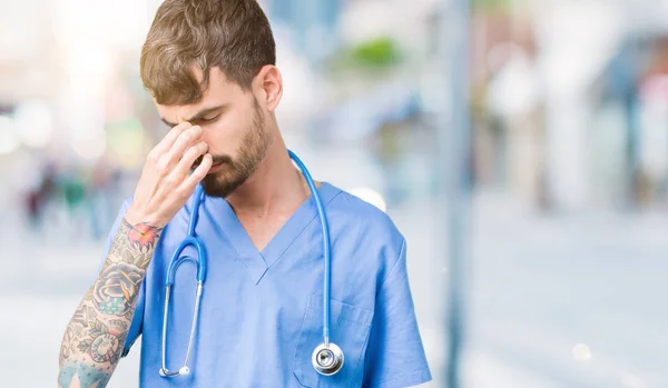 Young handsome nurse man wearing surgeon uniform over isolated background tired rubbing nose and eyes feeling fatigue and headache. Stress and frustration concept.
