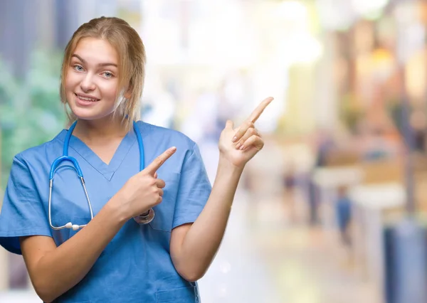 笑顔で側に つの手と指で指しているカメラを見て孤立の背景の上の外科医の制服を着て若い白人医師女性 — ストック写真