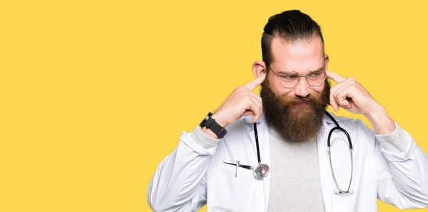 Young blond doctor man with beard wearing medical coat covering ears with fingers with annoyed expression for the noise of loud music. Deaf concept.