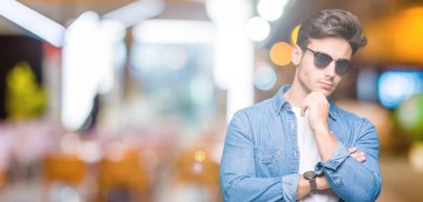 Genç yakışıklı adam çene düşünme hakkında soru, dalgın ifade üzerine el ile izole arka plan üzerinde güneş gözlüğü takıyor. Düşünceli yüzüyle gülümsüyor. Şüphe kavramı.