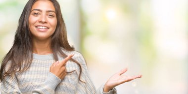 Hayret ve el ile sunulması ve parmak ile işaret ederken kameraya gülümseyen izole arka plan üzerinde kış kazak giyen genç güzel Arap kadın.