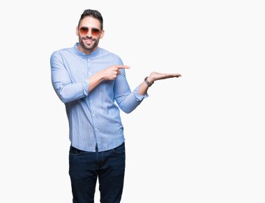 Yakışıklı delikanlı hayrete ve el ile sunulması ve parmak ile işaret ederken kameraya gülümseyen izole arka plan üzerinde güneş gözlüğü takıyor.