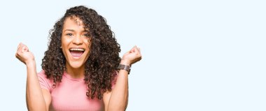 Kıvırcık saçlı pembe kutluyor t-shirt giyen genç güzel kadın şaşırttı ve başarı için kaldırdı kollarını açarak hayret ve gözleri açık. Kazanan kavramı.