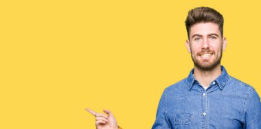 Kameraya bakarak yan el ve parmak ile işaret yüzünde, kocaman bir gülümseme ile rahat kot gömlek giymiş genç sarışın yakışıklı.