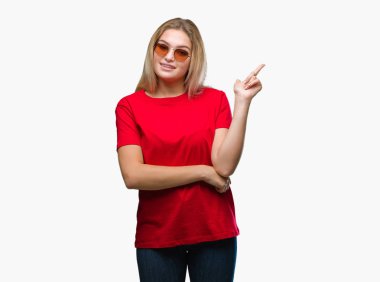 Kameraya bakarak yan el ve parmak ile işaret eden genç kafkas kadın yüzünde kocaman bir gülümseme ile izole arka plan üzerinde güneş gözlüğü takıyor.