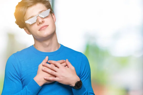 Young man wearing funny thug life glasses over isolated background smiling with hands on chest with closed eyes and grateful gesture on face. Health concept.