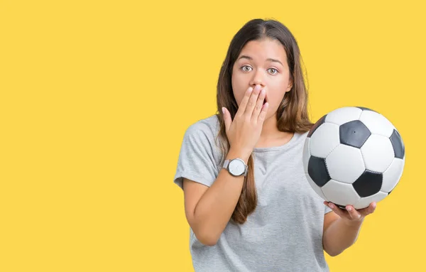 Young beautiful brunette woman holding soccer football ball over isolated background cover mouth with hand shocked with shame for mistake, expression of fear, scared in silence, secret concept