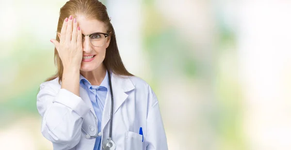 顔と驚きの感情に自信を持って笑顔で手で片目を覆う孤立の背景に医療のコートを着て中年中高年医師女性 — ストック写真