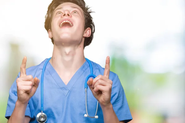 年轻的医生穿着医疗制服在孤立的背景惊讶和惊讶地抬起头来 用手指和举起的手臂指指点点 — 图库照片