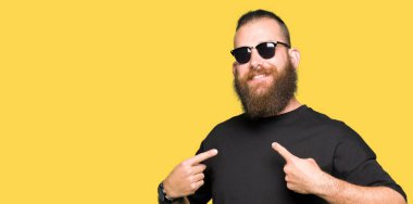 Yüzünde gülümseme ile kendinden emin görünüyorsun, kendini gururlu ve mutlu parmakları ile işaret güneş gözlüğü takmış genç hipster adam.