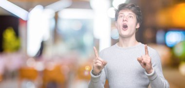 Yakışıklı delikanlı izole arka plan üzerinde kış kazak giyen hayrete ve yukarıya bakıyor ve parmakları ile işaret ve silah kaldırdı şaşırttı.