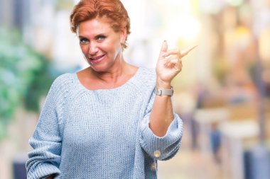 Kameraya bakarak yan el ve parmak ile işaret yüzünde kocaman bir gülümseme ile izole arka plan üzerinde kış kazak giyen Atrractive üst düzey beyaz Kızıl saçlı kadın.