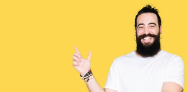 Uzun saç ve sakal, yüzünde kocaman bir gülümseme ile rahat beyaz t-shirt giyen genç hipster adam kameraya bakarak yan el ve parmak ile işaret.