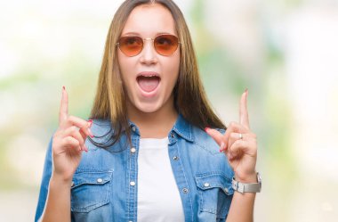 Genç güzel beyaz kadın hayrete ve yukarıya bakıyor ve parmakları ile işaret ve silah kaldırdı sürpriz izole arka plan üzerinde güneş gözlüğü takıyor.