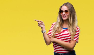 Kameraya bakarak yan el ve parmak ile işaret yüzünde kocaman bir gülümseme ile izole arka plan üzerinde güneş gözlüğü takmış genç güzel sarışın kadın.