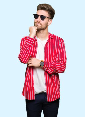 Genç yakışıklı adam çene düşünme hakkında soru, dalgın ifade üzerine el ile güneş gözlüğü takıyor. Düşünceli yüzüyle gülümsüyor. Şüphe kavramı.