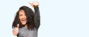 Kıvırcık saçlı onaylama gri kazak giyen genç güzel kadın el ile olumlu hareketi yapıyor gülümseyen ve başarı için mutlu yaşasın. Kamera, kazanan jest arıyorsunuz.