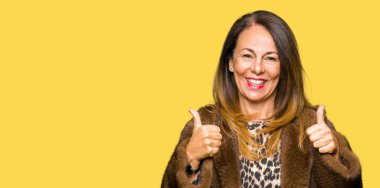 Vizon ceket başarı işareti el ile pozitif jest yapıyor giyen güzel orta yaş zarif kadın başparmak gülümseyen ve mutlu. Neşeli bir ifadeyle, kazanan jest kameraya bakıyor.