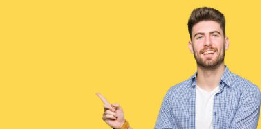 Kameraya bakarak yan el ve parmak ile işaret, yüzünde kocaman bir gülümseme ile rahat gömlek giymiş genç sarışın yakışıklı.