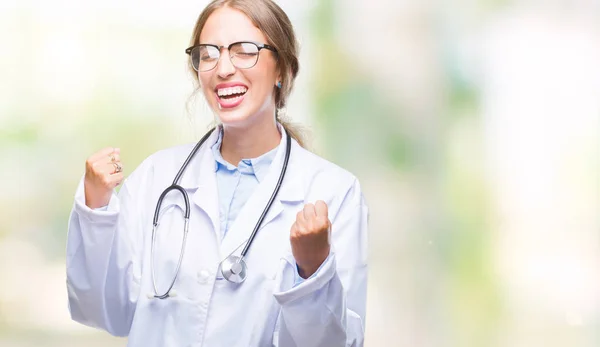 Beautiful young blonde doctor woman wearing medical uniform over isolated background very happy and excited doing winner gesture with arms raised, smiling and screaming for success. Celebration concept.