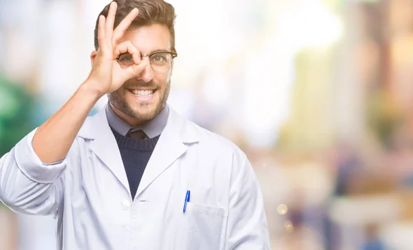 年轻英俊的男人穿着医生 科学家外套在孤立的背景做确定的手势与手微笑 眼睛通过手指看着快乐的脸 — 图库照片