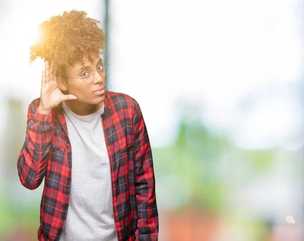 Beautiful young african american woman wearing glasses over isolated background smiling with hand over ear listening an hearing to rumor or gossip. Deafness concept.