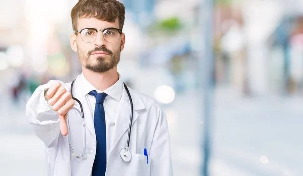 Young doctor man wearing hospital coat over isolated background looking unhappy and angry showing rejection and negative with thumbs down gesture. Bad expression.